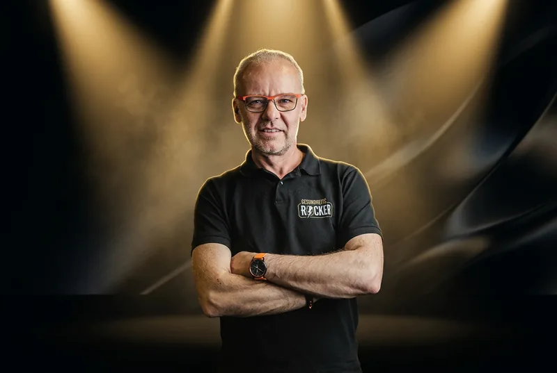 Jürgen Greis — Faszientherapeut, Neurospezialist und Gründer der Gesundheitsrocker. Verschränkte Arme, rote Brille, schwarzes Gesundheitsrocker-Polo, vor dramatischer Bühnenbeleuchtung mit goldenen Spotlights.